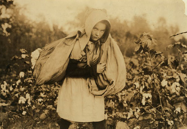 A young girl in a cotton field slightly hunched over as she carries a large bag across her shoulders. She peers slightly upward into the camera with a solemn expression.