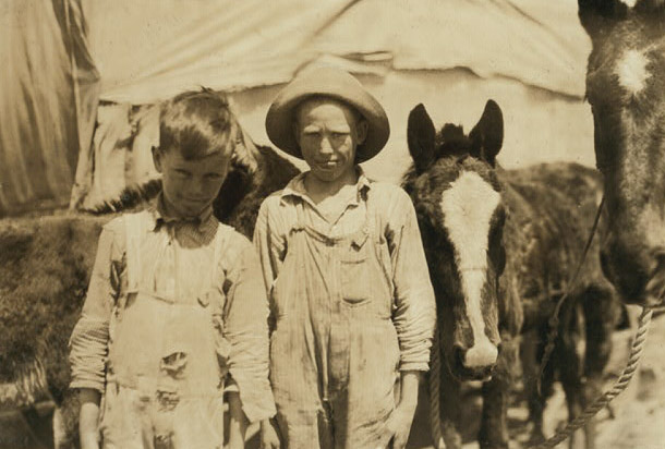 Two boys in ill-fitting, worn overalls look into the camera. Behind them are mules and a tent-like structure.