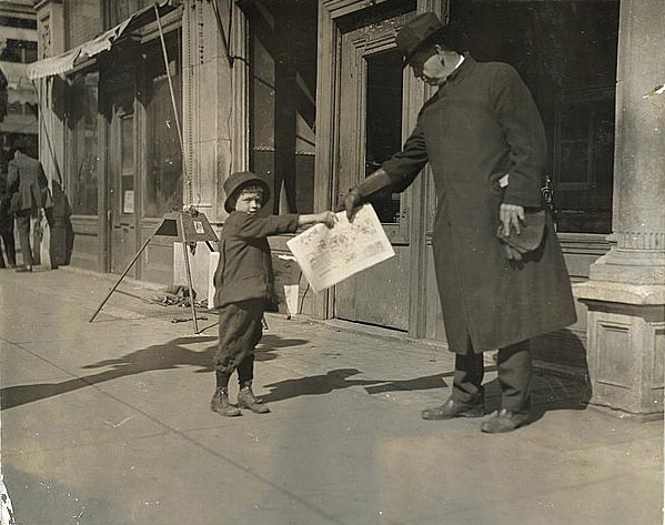 A young boy looks at the camera and hands a newspaper to a man. They stand on the sidewalk outside a business, and a few other people are in the background.