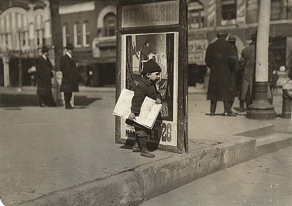 A very young boy stands alone on a sidewalk holding newspapers. The sign next to him reads 'Overholser' at the top. Multiple people and businesses are in the background. 
