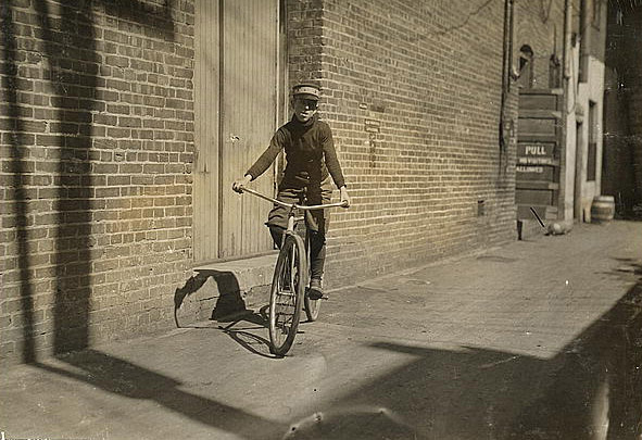 A boy rides a bike down a sidewalk. He looks directly into the camera. No other people are present.