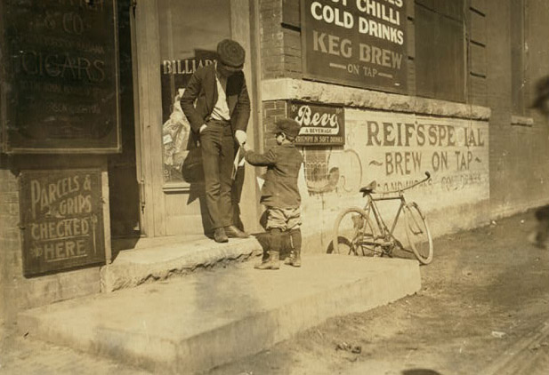 A small boy hands a newspaper to a man outside a business. There are advertisements for cigars, parcels, and beer. A small bicycle is parked nearby.