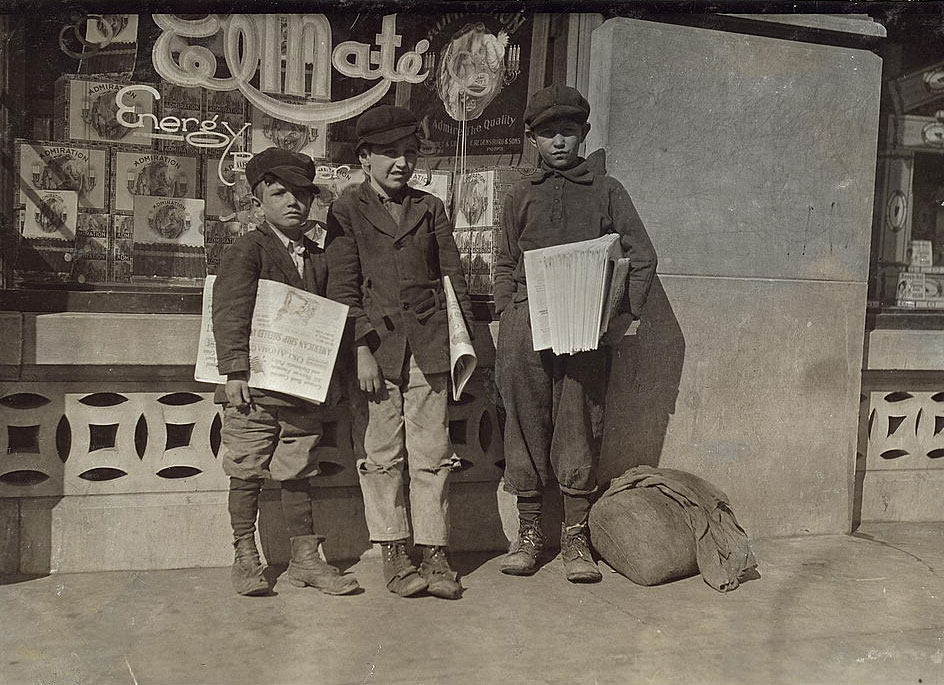 Three young boys stand outside a store front holding newspapers. Their clothes are worn and tattered. Boxes of cigars are seen through the store window. A large bulky bag lays at the feet of one of the boys.  