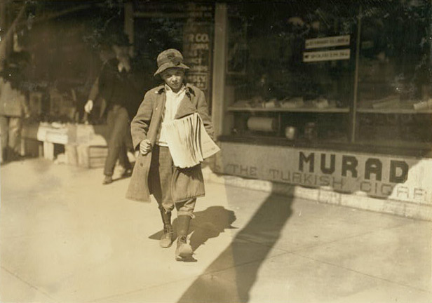 A young boy holding a handful of papers outside a storefront. The store has advertisements for Turkish cigars. A man in a suit and tie is walking out of the store.