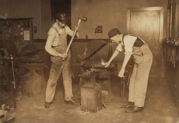 One young man raises a sledge hammer over an anvil while another holds something with a clamp on the anvil. Around them are hammers, anvils, and other large equipment. 