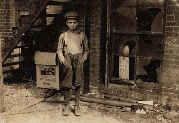 A boy stands outside a building with broken windows. He looks directly into the camera, his hands in his pockets. Suspenders hold up his too-large pants. 