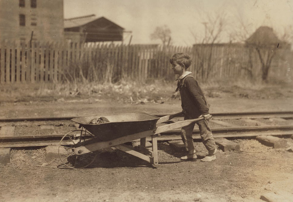 A young boy pushes a wheelbarrow along a railroad track. A wooden fence and a few structures are in the background