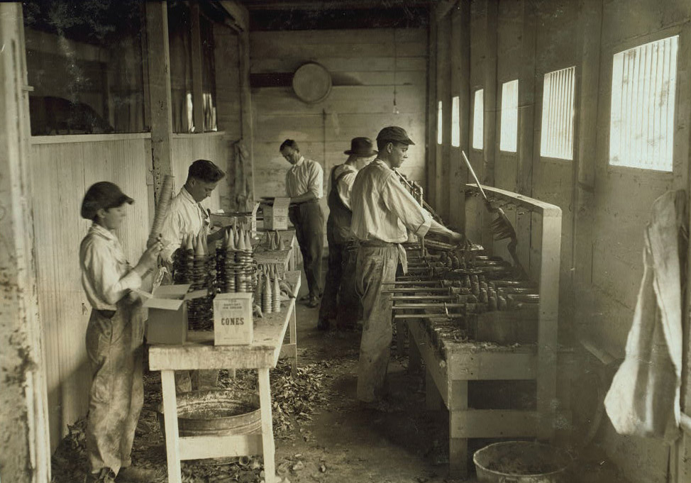 Four young men and a boy work in a dingy room. Two are stacking and packaging ice cream cones on a table. There are leaves and metal buckets on the floor.