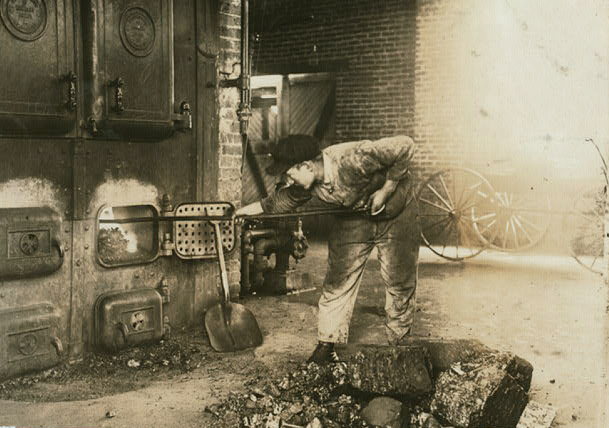 A boy in soot-stained clothes leans over pushing a lone fire-iron into a large furnace. A pile of ash and rubble is at his feet. 
