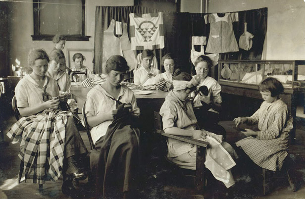 Several young women and look downward as they work on sewing projects. A small dress, quilt block, and other sewn pieces hang on the background. One woman in the back uses a sewing machine. 