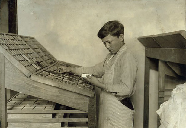 A boy in an apron focuses on pieces of metal type in large typesetters tray.