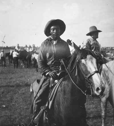 Bill Pickett in a cowboy hat sitting on his horse. Others standing and on horseback are visible in the background.