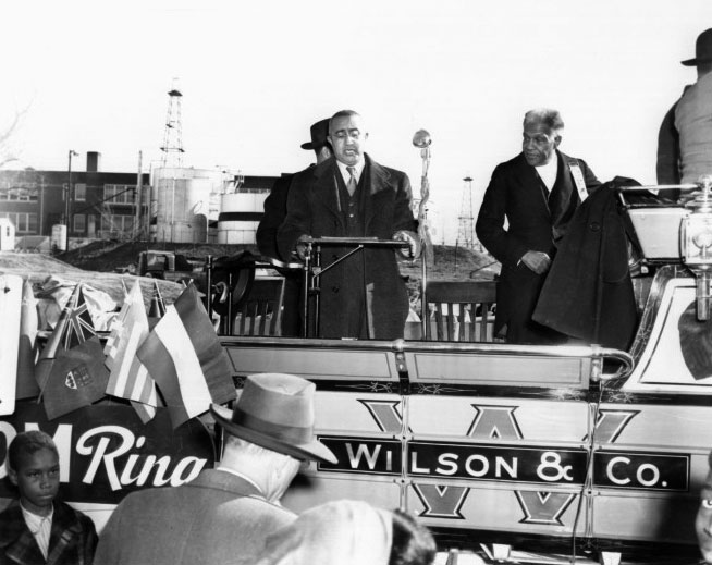Roscoe Dunjee stands on a raised platform, possibly a parade float, in front of a microphone. A few people can be seen standing at ground-level, and a display of multiple flags is visible.