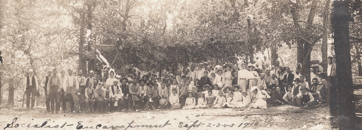 A group of about 80 people in a wooded area pose for a portrait. Behind them are several small tents and a log cabin. The handwritten text reads, 'Socialist Encampment Sept 2-3-4-5 1910.'