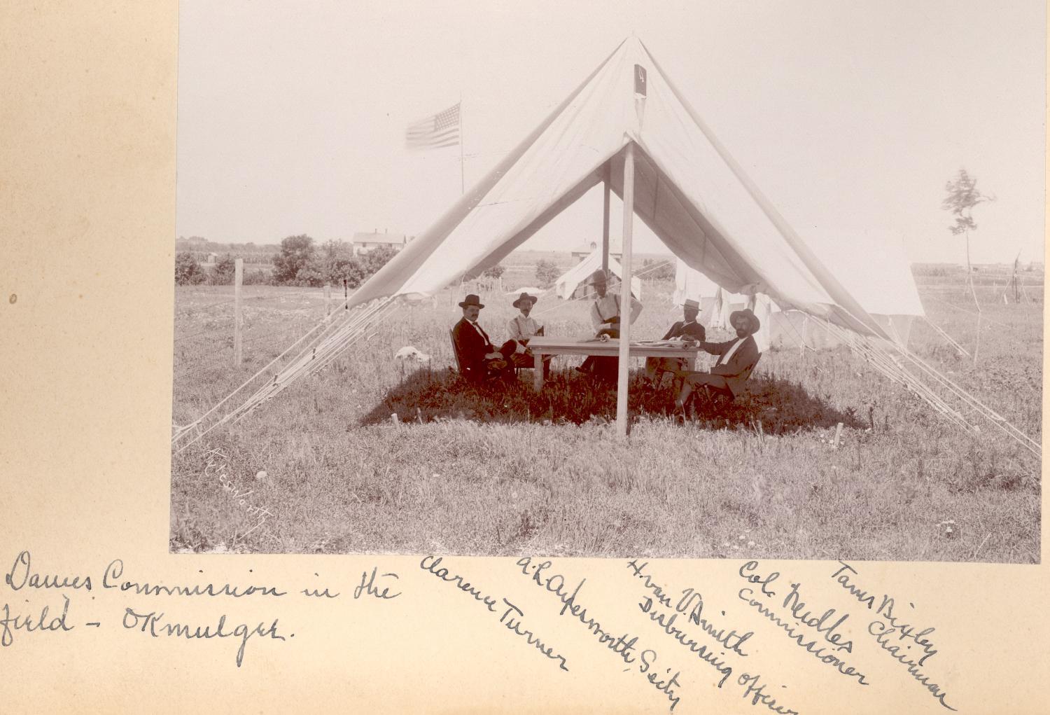Historic photograph of five men around a table inside a white tent pitched in an open field. The men wear suits and hats; two men wear white shirts and suspenders.