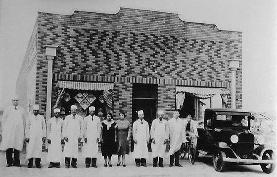 A historic photograph of a group of mostly men people standing outside a brick building next to a car. Most are wearing long aprons and hats. Two women wearing dresses stand with the group.