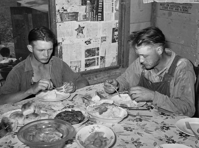 Sons of tenant farmer at noonday meal. Muskogee, Oklahoma in 1939. In this historic photograph, two men in worn clothing sit at a table eating. The walls of the room are covered in newspapers.