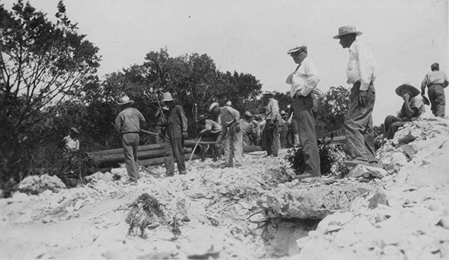 A group of men wearing hats are standing on uneven ground. Some have tools like shovels. 