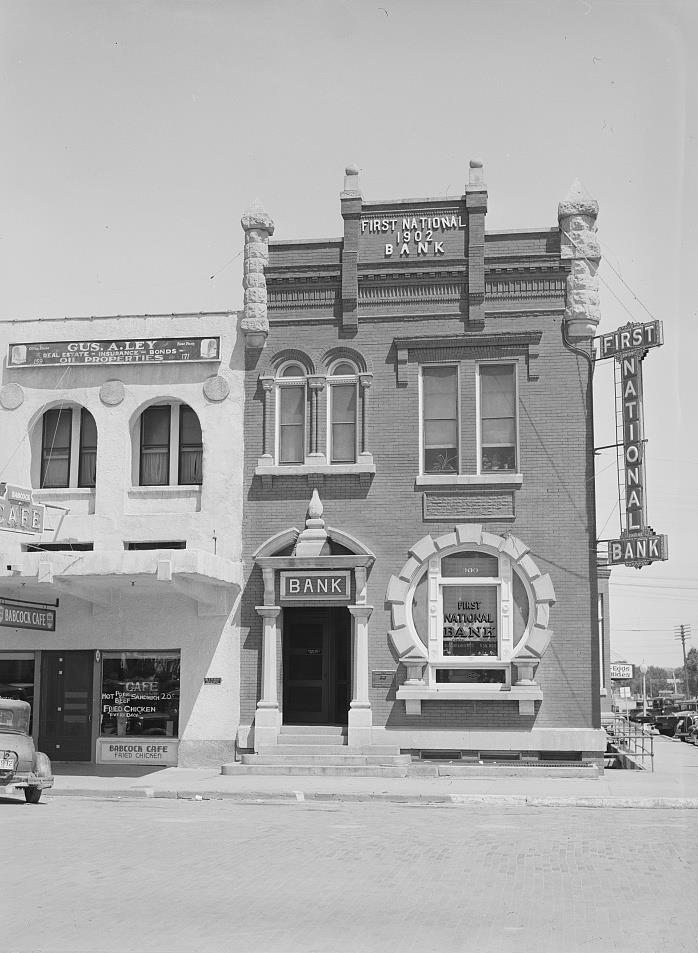 Historic photograph of building with multiple signs reading, First National Bank. The bank is a two-story structure connected to another building. 