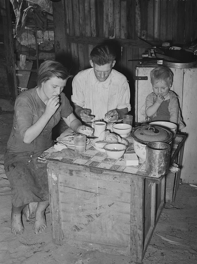 A woman, man, and toddler in tattered clothing sit around a crate on a dirt floor. They are eating from dishes on top of the crate.