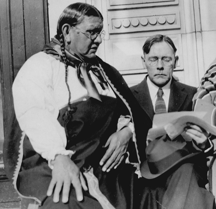 Chief White Eagle with this dark hair braids, wearing glasses and traditional Osage clothing stands with John Collier, a shorter man, in a modern suit. 