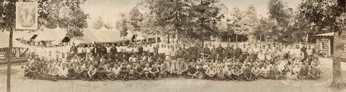 A panoramic photograph of about 150 men lined up for a picture in an outdoor area surrounded by trees and temporary tents. 
