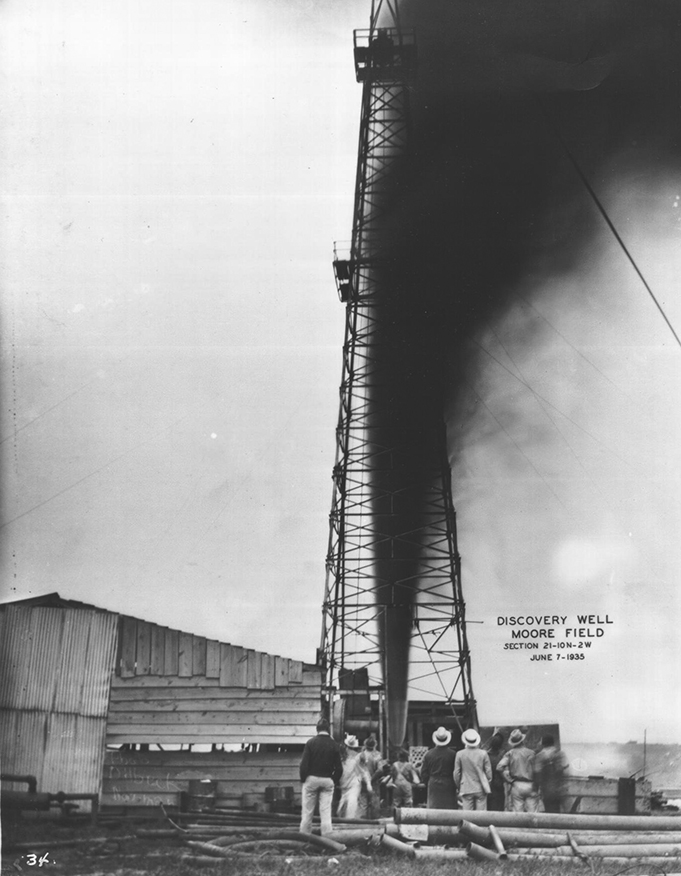A group of men look up at oil spewing out of an oil well. Text on photo says, Discovery Well Moore Field.