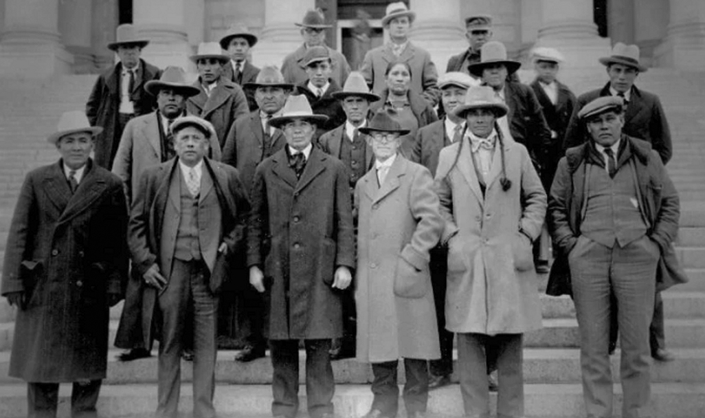 A group of about twenty men and one woman standing on step of a large building. All the men are wearing hats and many of the men are wearing overcoats. 