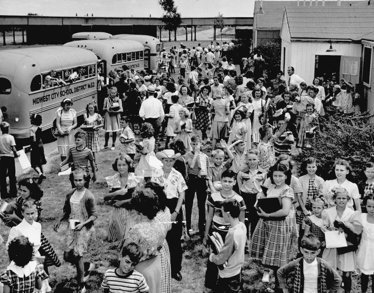 A large group of adolescents holding books standing outside of buses labeled 'Midwest City School District.'