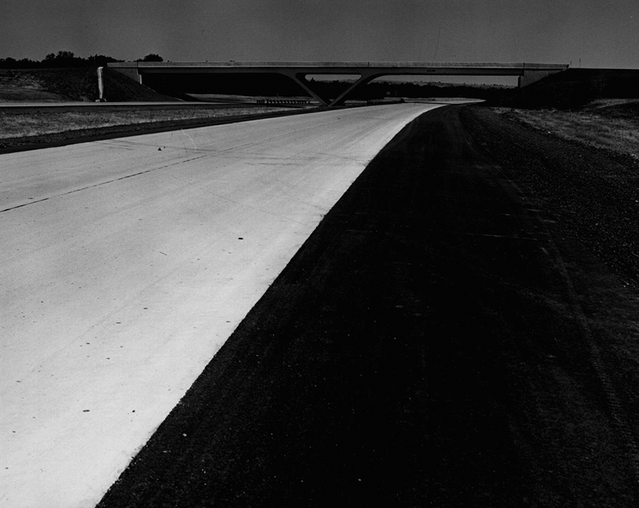 A long stretch of road contrasts with the land. There is a bridge in the background.  