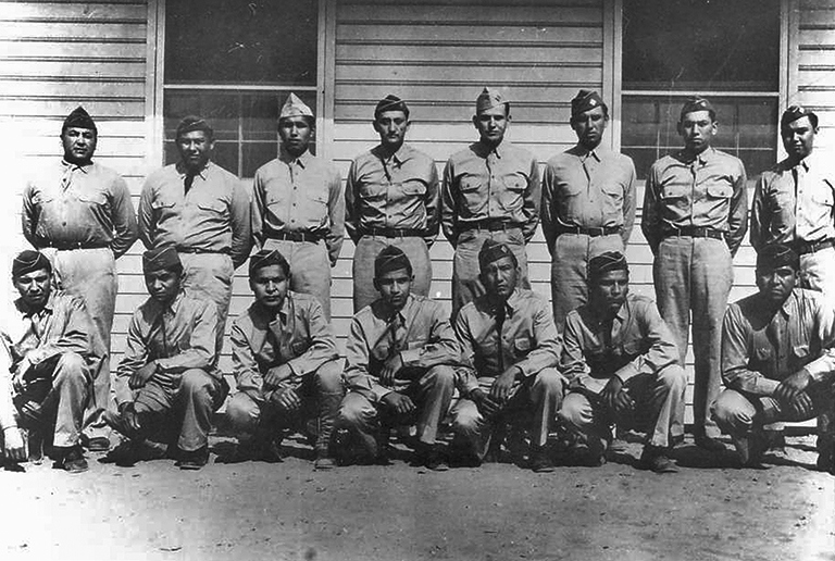 A group of 15 Comanche soldiers in uniform in front of a building.  Some stand and some kneel as pose for the photograph.