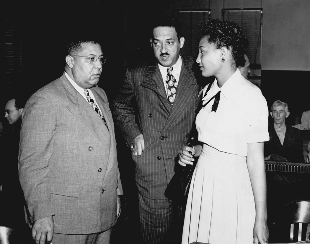 Two Black men in suits, Amos T. Hall and Thurgood Marshall, stand with Ada Louis Sipuel Fisher. They are inside a courtroom.
