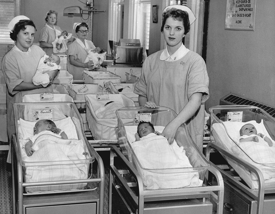Four nurses in uniform stand in a nursery. Three hold babies in their arms, and multiple babies lay in small beds.