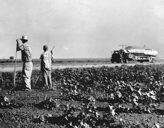 A man and a young boy in a field hold farming implements as they watch a large truck hauling a missile.