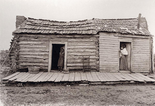 A log cabin with a wood roof. An empty rocking chair sits in front of the cabin. In one doorway stands a young woman. In another doorway stands a woman holding  a baby.