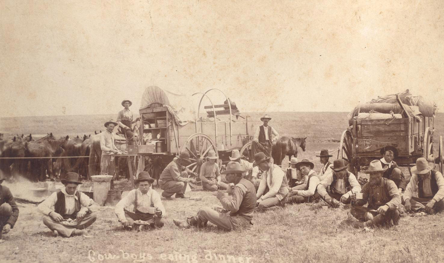 Twenty or so men sit and eat outdoors. Several horses are fenced off to one side. A chuckwagon and another wagon parked behind the group.