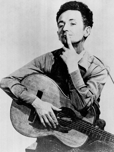 Historic photograph of Woody Guthrie, holding a finger to his lips with the other arm wrapped around a guitar.