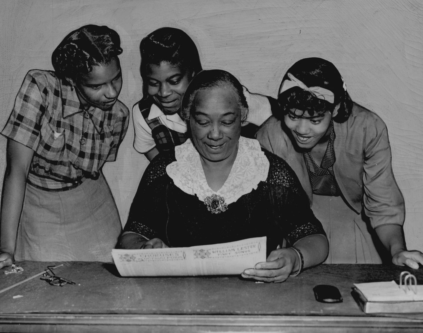A teacher sits at a desk surrounded by three students as they read a book of choruses together.