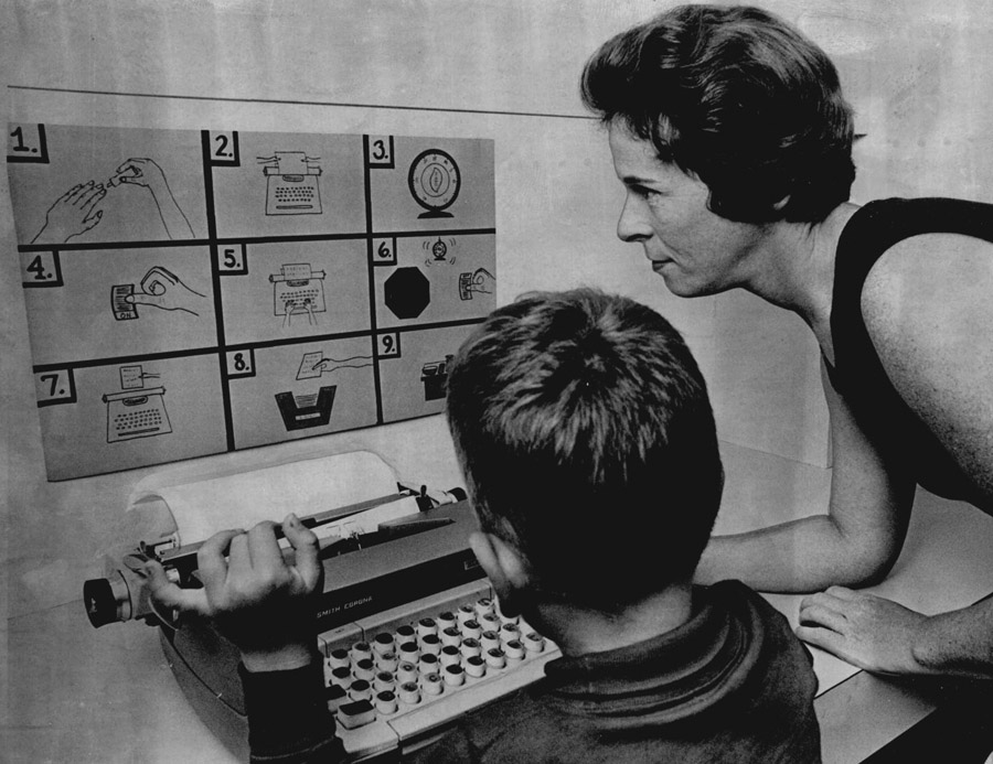 A young boy sits at a typewriter while a woman instructs him based on the images on the board in front of them.