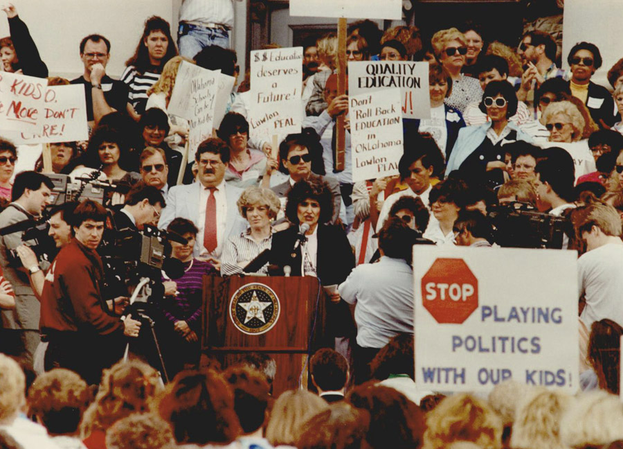 A woman speaks at a podium surrounded by demonstrators. Some hold signs with messages such as 'STOP PLAYING POLITICS WITH OUR KIDS!'