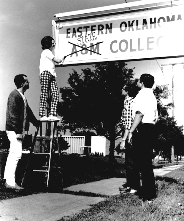 Three students look on as a young woman on a ladder crosses out A and M on a large sign. The word State is written in its place.