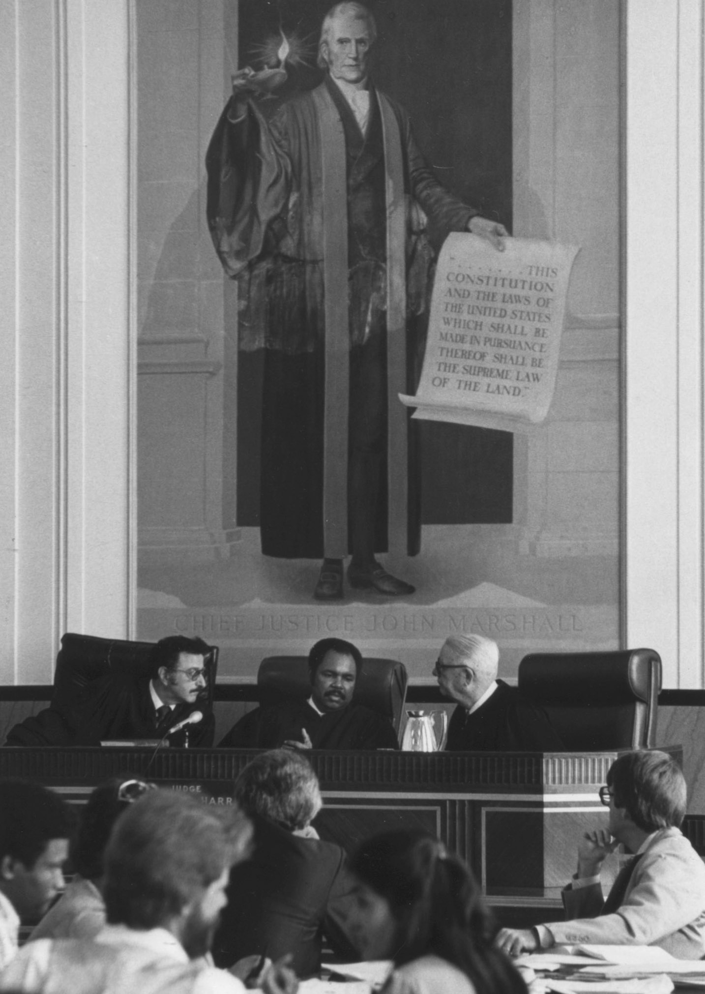 Three judges confer beneath a massive painting of former United States Chief Justice John Marshall, who holds a scroll of the Constitution. Court staff and clients sit in the foreground.