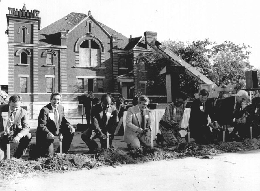 Eight people kneel to place stakes in the ground at a construction site in front of an historic church building with boarded windows.