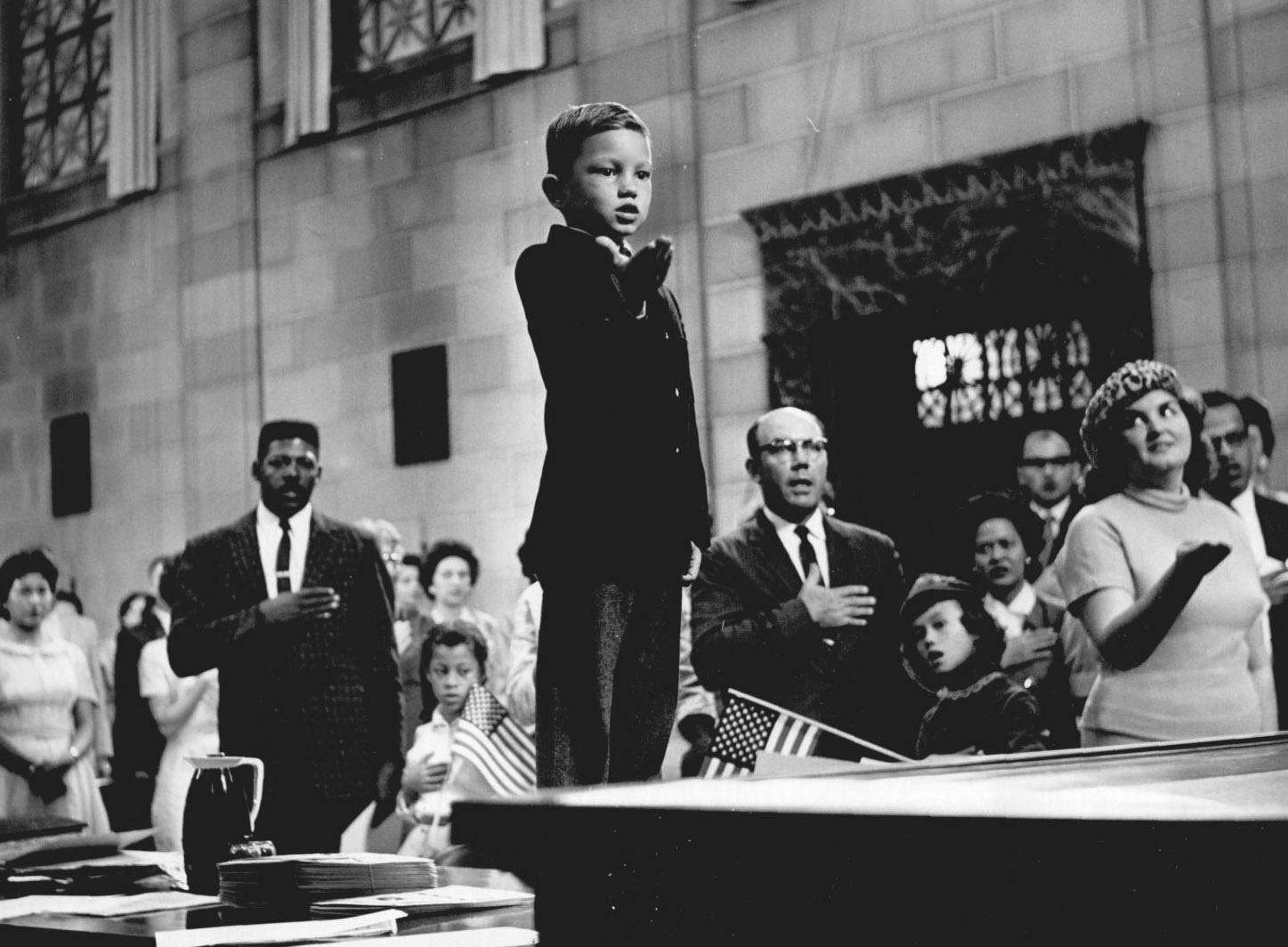 A child stands on a table facing the judge and flag at the front of the courtroom. He and the adults standing in the pews behind him hold their hands up facing forward or on their heart in allegiance.