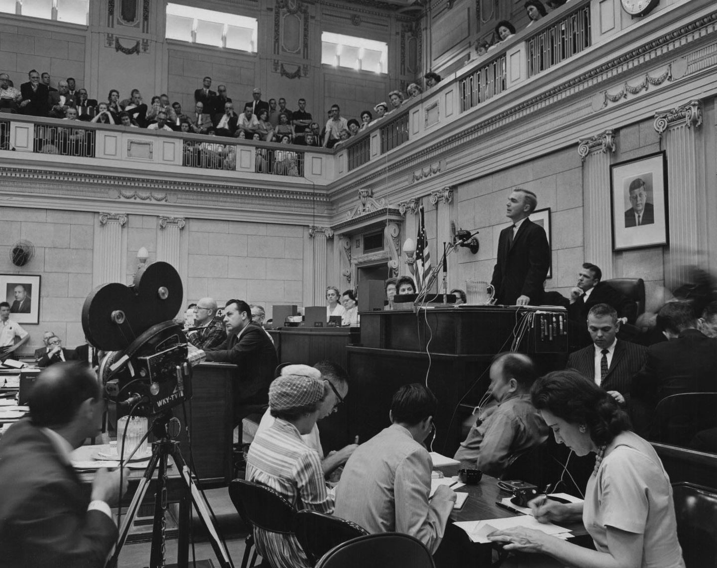 A speaker stands at the podium at the house or senate floor. Press and staff sit at desks all around, with a video camera at middle left, and many people observing from the second-floor gallery.