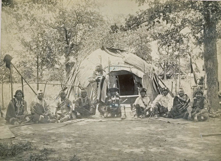 Nine people in traditional clothing and regalia sit on mats in front of a round bark lodge. The chief stands in the middle of the group wearing a blanket and a bear claw necklace.