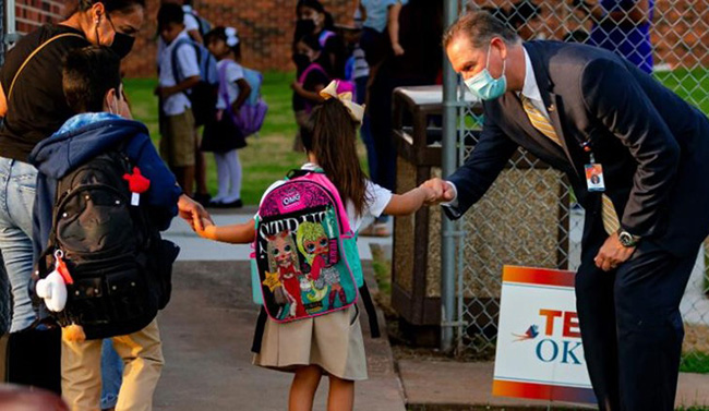 Man in suit greeting schoolchildren outside of a gate. 
