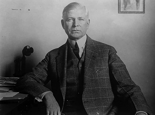 Elmer Thomas in a high white collar, tie, and plaid coat and vest sitting in front of a mostly bare wall except for a small photo in the upper right corner. 