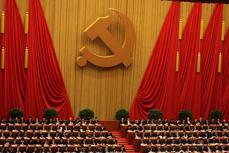 A large chamber with about one hundred people seated at desks before a large wall adorned with red and gold drapes and a hammer and sickle