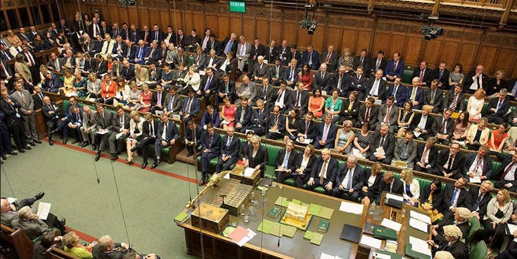 Many people seated in a small chamber. In the center of the chamber is a large table, and the people at the table are wearing white wigs. 